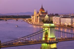 Vedere nocturnă asupra Budapestei, cu Podul cu Lanțuri iluminat în prim-plan și Clădirea Parlamentului Ungariei în fundal, reflectând frumusețea istorică și arhitecturală a orașului de-a lungul râului Dunărea